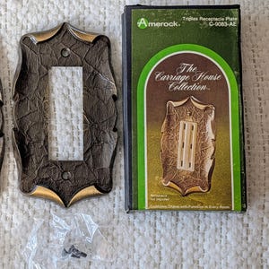 May include: Decorative brown and gold triplex receptacle plates from The Carriage House Collection. The plates have a textured finish and a decorative border. The packaging includes the Amerock brand name and product information.