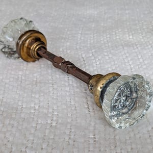May include: A pair of clear, faceted glass door knobs connected by a central metal spindle. Each knob features a circular, multi-faceted design with a brass-toned metal base that attaches to the threaded square metal rod connecting the two pieces.