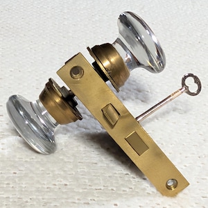 May include: A vintage door lock set featuring clear glass doorknobs, brass hardware, and a small key. The lock mechanism is rectangular with a keyhole and a sliding bolt. The doorknobs are oval-shaped.