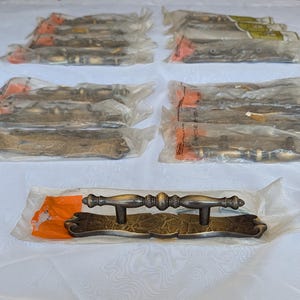 May include: A collection of nine metal drawer pulls, each individually packaged in clear plastic. The pulls feature a dark bronze finish with a textured, crackled pattern on the rectangular backplate and a central handle with decorative detailing.