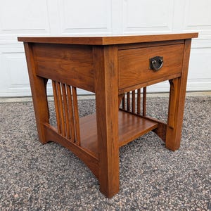 May include: A square wooden end table with a medium brown finish, featuring a single drawer with a dark metal drop pull handle, four straight legs, and a lower shelf supported by vertical slats on the sides of the furniture piece.