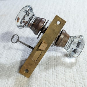May include: A door lock mechanism featuring two clear, faceted glass knobs attached to a rectangular brass faceplate. The assembly includes a metal key inserted into the side of the brass plate, which also features two rectangular latch components.