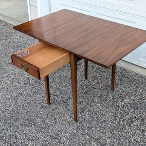 May include: A brown wooden drop-leaf table with four tapered legs and a single pull-out drawer featuring a brass handle. The interior of the drawer displays a label with the text SOLID MAPLE AND CHERRY. The table surface is rectangular.