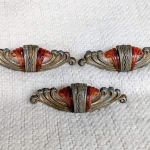 May include: Three metal drawer pulls featuring a central vertical panel with leaf-like carvings, flanked by curved, fan-shaped wings. The metal has a weathered, muted finish, while the central accents are a translucent, mottled amber-brown color.