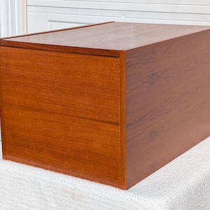 May include: A rectangular wooden storage unit with a warm brown finish and visible wood grain. The piece features a flat top surface and a single front panel that appears to be a drawer or door, showcasing a clean, boxy construction.
