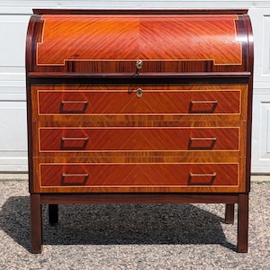 May include: A wooden roll-top desk featuring a curved tambour door with a diagonal wood grain pattern, three horizontal drawers with thin metal handles, and a dark brown frame. The desk includes two brass keyholes centered on the front panels.