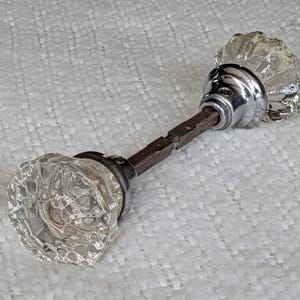 May include: A door knob set featuring two clear, faceted glass knobs with a multi-sided geometric shape. Each knob is attached to a polished chrome-colored metal base, connected by a central, square-shaped, dark brown metal spindle rod.