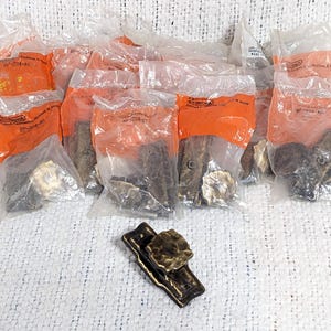 May include: A close-up of a package of antique brass cabinet hardware. The hardware is in individual plastic bags with orange labels. The hardware is in the shape of a small, ornate knob.
