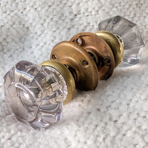 May include: A collection of vintage door knobs. The knobs feature clear glass and brass elements. The glass knobs have a faceted and floral design. The brass components show signs of age and wear. The knobs are arranged in a row.