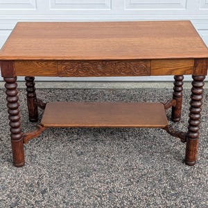 May include: A rectangular wooden table with a medium brown finish, featuring a flat top surface and a lower shelf. The table has four legs with a turned, spool-style design and a central drawer front decorated with an intricate carved floral pattern.