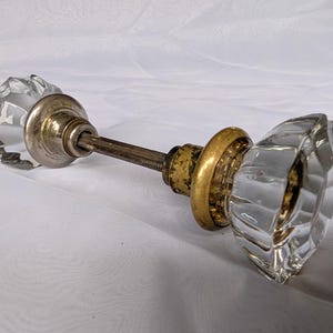 May include: A door knob set featuring two clear, faceted glass knobs connected by a central metal spindle. Each knob is attached to a circular metal base, with one base showing a tarnished silver tone and the other a worn, aged brass finish.