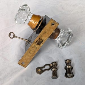 May include: A mortise lock set featuring two clear glass knobs with brass collars, a rectangular brass faceplate, and a metal skeleton key inserted into the mechanism. Two separate metal keyhole escutcheons are positioned below the lock assembly.