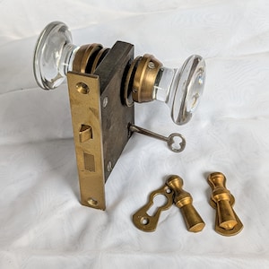 May include: A mortise door lock set featuring two clear glass knobs with brass bases, a rectangular brass faceplate, and a small metal key. Included are two separate brass escutcheon plates with matching brass covers for keyhole protection.