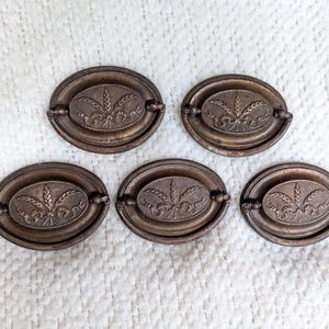 May include: Five oval-shaped brass drawer pulls featuring a central embossed design of wheat stalks tied with a ribbon. Each pull has a matching brass backplate and a hinged drop handle, showcasing a weathered, aged metal finish with dark patina.
