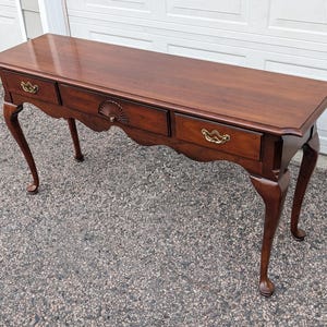 May include: A mahogany-colored console table with three drawers and brass hardware. The table has a scalloped apron and curved legs. The drawers have decorative pulls, and the center drawer features a fan-shaped design.