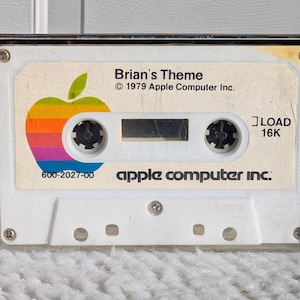 May include: A vintage audio cassette tape with a white label featuring the Apple Computer Inc. logo, a colorful rainbow apple. The label reads "Brian's Theme" and "© 1979 Apple Computer Inc.". The cassette is in a clear plastic case.