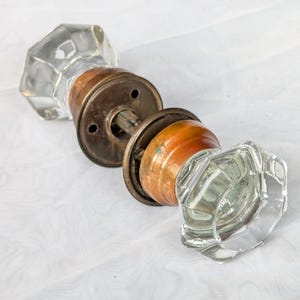 May include: A vintage door knob set featuring two clear, multi-faceted glass knobs attached to circular, aged copper-toned metal rosettes. The two knobs are connected by a central metal spindle, showcasing a classic hardware design with visible patina.
