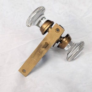 May include: A door latch assembly featuring two clear glass knobs with a smooth, rounded profile. The knobs are connected by a central brass-toned metal plate that includes a rectangular latch bolt and two circular screw holes for mounting.
