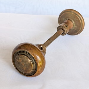 May include: A vintage brass door knob set featuring a spherical handle with a weathered patina, connected by a square metal spindle to a circular mounting plate. The hardware displays visible signs of age and oxidation on the metallic surface.