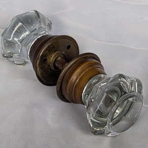May include: A door knob set featuring two clear, faceted glass knobs with an octagonal shape. The knobs are connected by a central metal spindle and include two circular, weathered brass-toned mounting plates with visible screw holes.