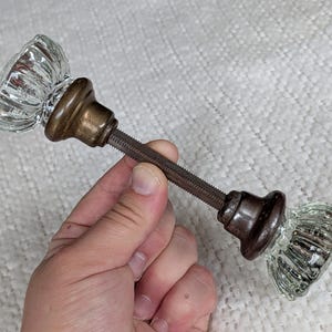 May include: A pair of clear, fluted glass door knobs connected by a long, threaded metal spindle. Each knob features a rounded, ribbed design with a dark, aged bronze-toned metal base that secures the glass to the central connecting rod.