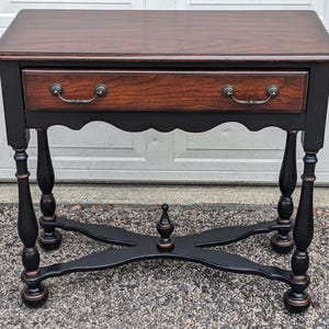 May include: A dark wood side table with a single drawer and ornate legs. The tabletop and drawer front are a rich brown, while the frame and legs are black with bronze accents. The table has a classic, traditional design.