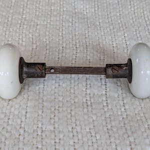May include: A vintage door knob set featuring two smooth, off-white porcelain knobs connected by a dark, weathered metal spindle. The metal components include small screws for securing the knobs to the central rod, showing signs of age and wear.
