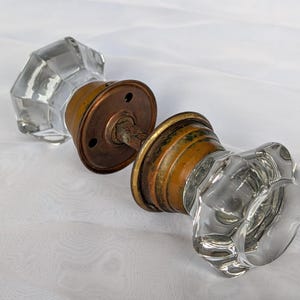 May include: A pair of clear, faceted glass door knobs connected by a central metal spindle. Each knob features a circular, weathered brass-toned metal base plate with visible screw holes, showing signs of oxidation and age-related patina.