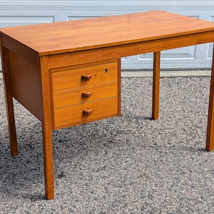 May include: A wooden desk with a warm, reddish-brown finish. The desk features three drawers on the left side with curved wooden handles. The desk has four slender legs and a rectangular tabletop. The desk measures approximately 48 inches long.