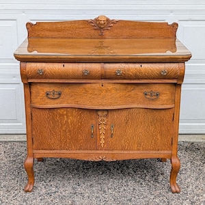 May include: A medium-brown oak wood sideboard featuring a top backsplash with a carved lion head motif, two upper drawers with metal pulls, one wide lower drawer, and two cabinet doors with central floral carvings, supported by four curved legs.