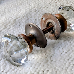 May include: Two vintage clear glass door knobs with faceted designs. Each knob has a metal base and a connecting rod. The metal components show signs of age and wear, adding to their antique charm.