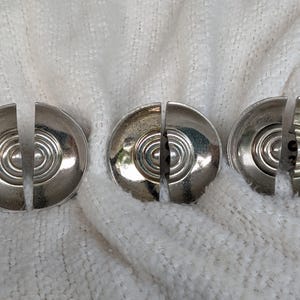 May include: Three circular silver-toned metal buttons, each featuring a split design with concentric circular ridges radiating from a central point. The buttons have a polished, reflective finish and are arranged in a horizontal row.