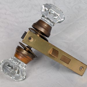 May include: A door handle set featuring two clear, faceted glass knobs attached to a brass-toned metal plate. The mechanism includes a rectangular brass latch plate with two visible openings and a circular screw hole at the end of the metal arm.