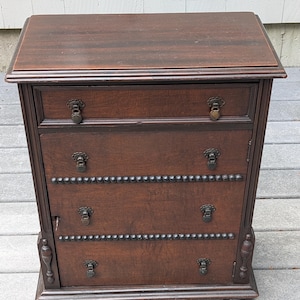 May include: A dark brown wooden chest of drawers featuring a rectangular top, one upper drawer, and a lower cabinet section with two doors. The cabinet doors are adorned with horizontal rows of decorative studs and small metal drop handles.