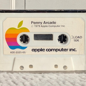 May include: A vintage Apple Computer Inc. cassette tape for "Penny Arcade" from 1979. The tape features the classic rainbow Apple logo and the text "LOAD 16K". The cassette is in a clear plastic case.