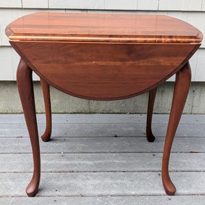 May include: A brown wooden drop-leaf table with a smooth rectangular top and two semi-circular hinged side panels. The table features four curved cabriole legs that extend outward from the base, showcasing a rich, polished wood grain finish.