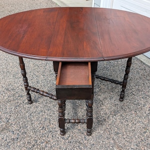 May include: A dark brown, oval-shaped antique table with a drawer. The table has a wooden surface and turned legs. The drawer is open, revealing the interior. The table is set on a gray surface.