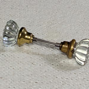 May include: A pair of vintage glass door knobs with brass hardware. The clear glass knobs have a ribbed design, and the brass components show signs of age. The knobs are connected by a metal rod, suggesting they are ready for installation.