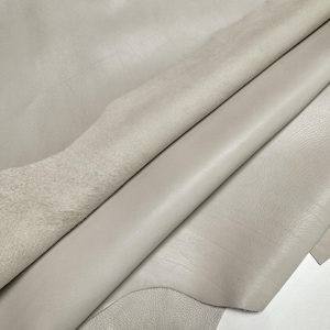 May include: A large piece of light gray leather folded over to reveal both the smooth, finished top grain surface and the soft, textured suede underside. The material displays natural folds and creases across its surface.