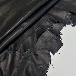 May include: A large piece of black leather with a smooth, matte finish and a natural, irregular edge. The material is folded over itself, creating deep, soft creases and shadows across the surface, highlighting the texture of the hide.