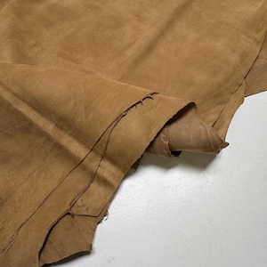 May include: A large, irregular piece of tan suede leather with a soft, matte texture. The material features natural edges and a folded section that reveals the underside, showcasing the fibrous, unfinished texture of the hide's reverse side.