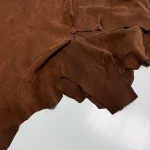 Pode incluir: Uma peça grande de camurça castanha de forma irregular com uma textura macia e mate. O material está drapeado em dobras, mostrando a sua natureza flexível e a cor castanha profunda e consistente em toda a superfície da pele.