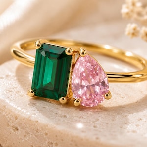 May include: A gold ring featuring two prominent gemstones: a rectangular emerald-cut green stone and a pear-shaped pink stone. The stones are set side-by-side on a polished gold band, creating a distinct contrast between the vibrant green and soft pink hues.