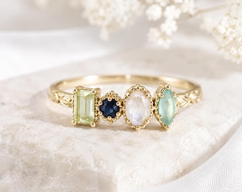 Peut inclure: Une bague en or ornée de quatre pierres précieuses distinctes alignées : une pierre rectangulaire vert clair, une petite pierre ronde bleu foncé, une pierre ovale blanc irisé et une pierre vert clair en forme de marquise sur un anneau texturé.