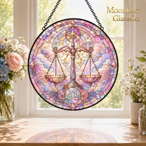 Könnte beinhalten: Ein runder Buntglas-Sonnenfänger mit dem Waage-Tierkreiszeichen in Rosa-, Lila- und Blautönen. Das Design zeigt eine zentrale Waage, ein Sternmotiv und Wolken, wobei der Text Moonleaf GlassCo. in der oberen rechten Ecke zu sehen ist.