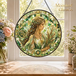 Op de afbeelding: Een ronde glas-in-lood zonnevanger met een vrouw die tarwe vasthoudt, als symbool voor het sterrenbeeld Maagd. Het ontwerp bevat groene en gouden glassegmenten, een zwart metalen frame en een ketting. De tekst luidt Moonleaf GlassCo.