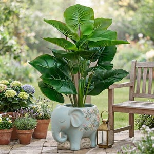 May include: A light blue ceramic elephant planter featuring a floral design on its side and the text "for someone cuter" printed on its base. The planter holds a large artificial green plant with broad, veined leaves and sturdy stems.