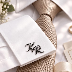 Op de afbeelding: Een witte overhemdmanchet met een zwarte metalen manchetknop met de letters KR in cursief schrift, gecombineerd met een gestructureerde beige das. Het overhemd heeft een strakke witte kraag en een gladde afwerking.