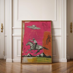 May include: A framed art print featuring a vibrant hot pink background with a black and white illustration of a cowboy riding a horse while being abducted by a flying saucer. The scene includes mountain silhouettes and an orange rectangular accent.
