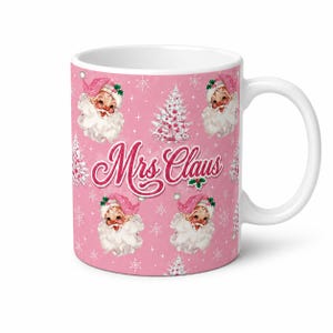 May include: A white ceramic mug featuring a pink background with a repeating pattern of vintage-style Mrs. Claus faces wearing pink hats, white Christmas trees, and snowflakes. The text Mrs. Claus is printed in a red cursive font in the centre.